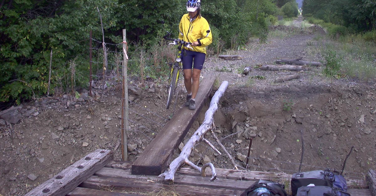 Beate Steger trägt ihr Rad über eine wackelige Holzkonstruktion auf die teilzerstörte Eisenbahnbrücke